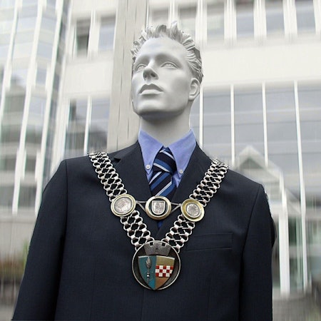 Eine Puppe trägt die Amtskette des Bürgermeisters vor einem Rathaus.