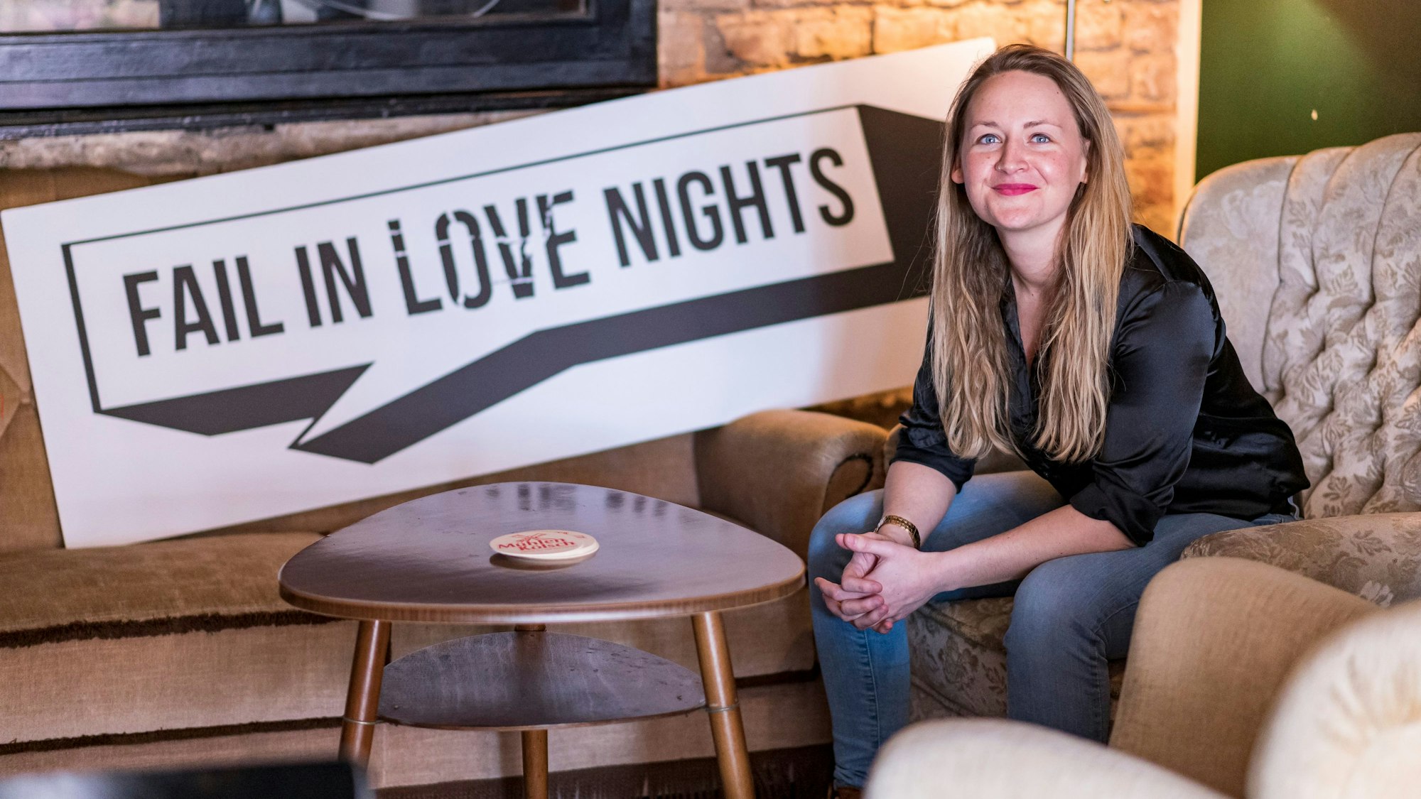 Juliana Stockheim sitzt auf einem Sofa in einer Bar. Neben ihr ein Schild auf dem „Fail in Love Nights“ steht.