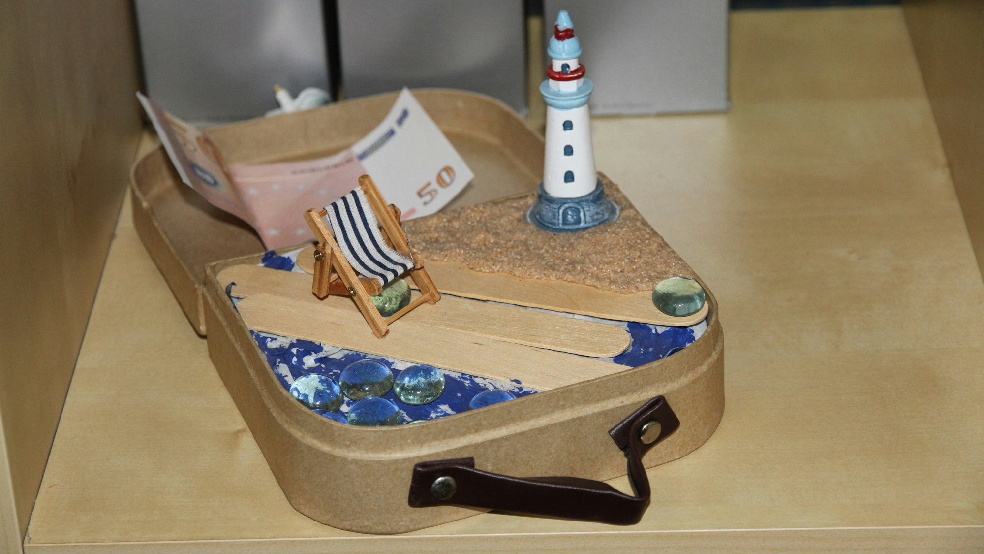 Ein selbstgebasteltes Reiseköfferchen mit einem Leuchtturm am Meer, einem Liegestuhl und einem Geldschein