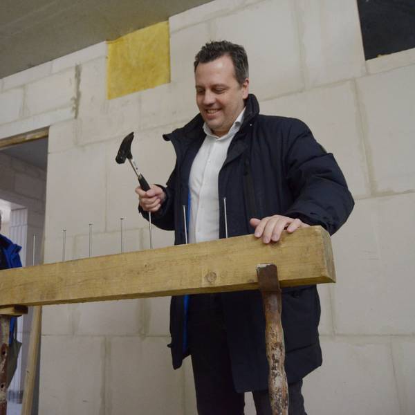 Sacha Reichelt treibt mit einem Hammer einen von sieben Nägeln in einen Balken. Mehrere Zuschauer im Hintergrund lachen.