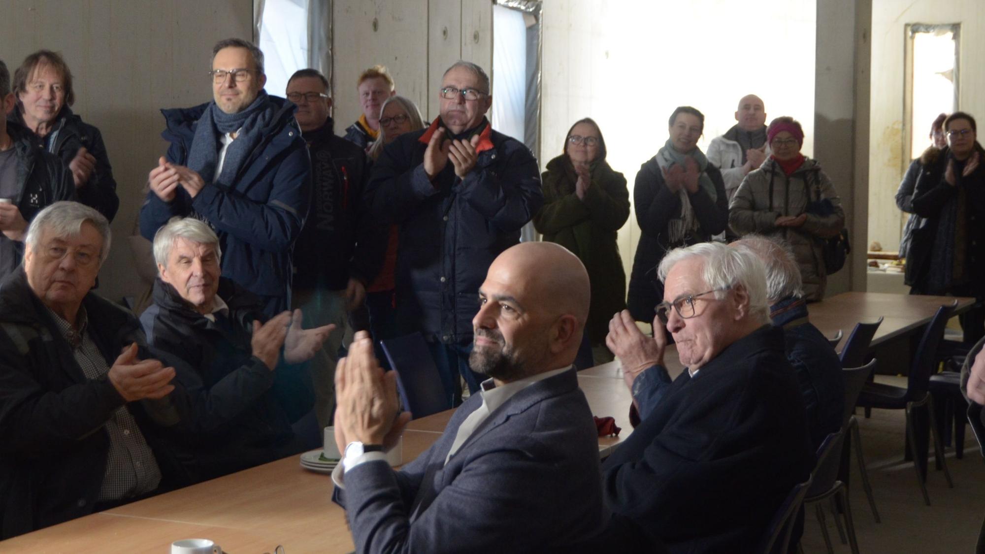 Besucher und Besucherinnen des Richtfestes sitzen und stehen in einem der Räume im Erdgeschoss und klatschen Beifall.