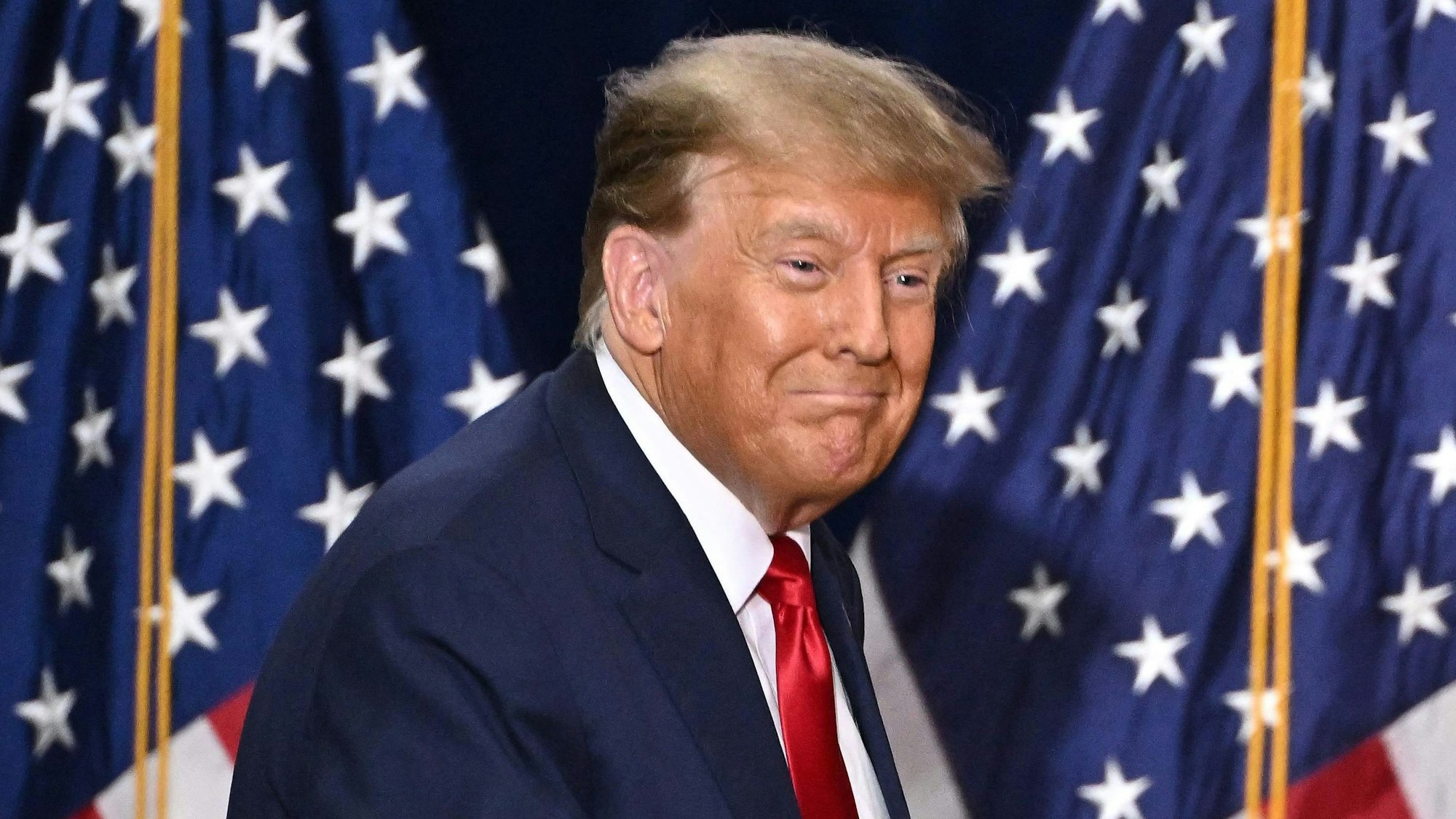 Ex-US-Präsident Donald Trump steht auf einer Bühne vor zwei Flaggen der Vereinigten Staaten und blickt lächelnd ins Publikum.