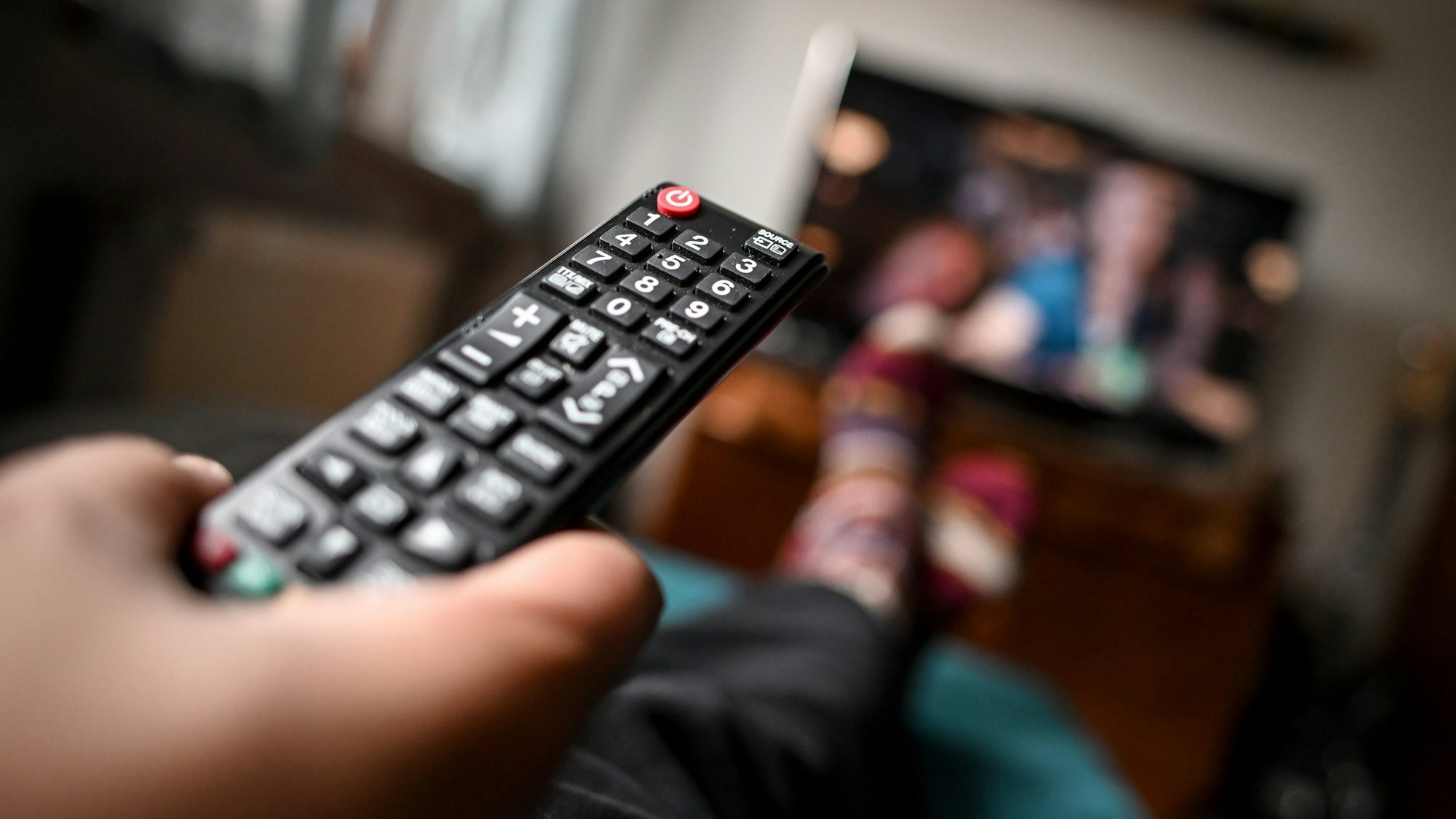 Eine Person liegt auf einem Sofa vor einem Fernseher und hält eine Fernbedienung in der Hand