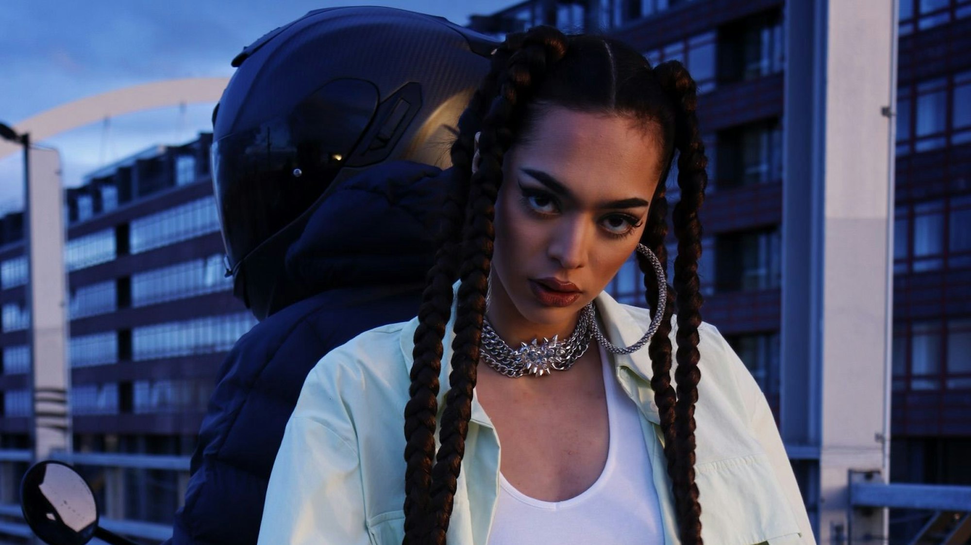 Eine Frau mit langen Zöpfen sitzt Rücken an Rücken mit einem behelmten Motorradfahrer. Im Hintergrund sind mehrstöckige Gebäude zu sehen.