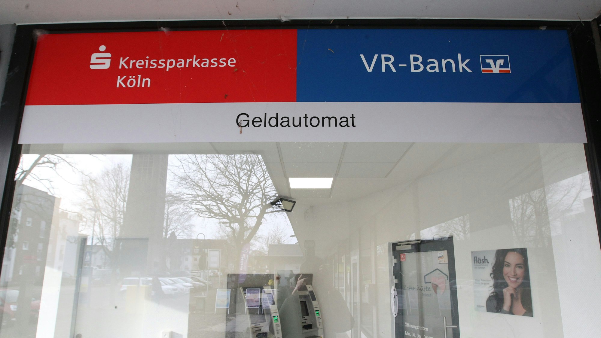 Blick in den Selbstbedienungsraum einer Bankfiliale. Auf der Scheibe kleben die Logos und Schriftzüge Kreissparkasse Köln und VR-Bank.