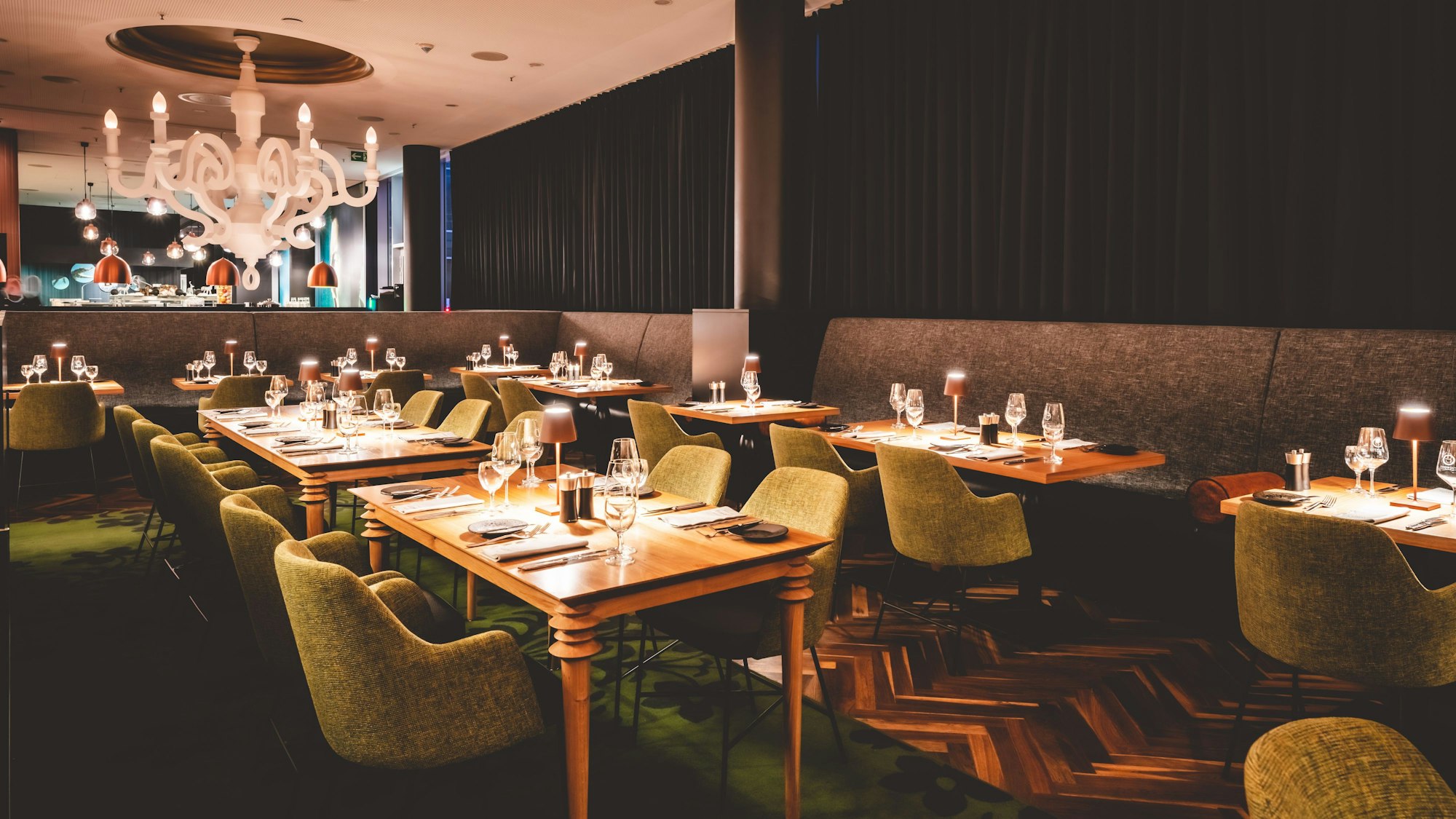 Furnierte Holztische und grüne Sessel im Innenraum des Restaurants.