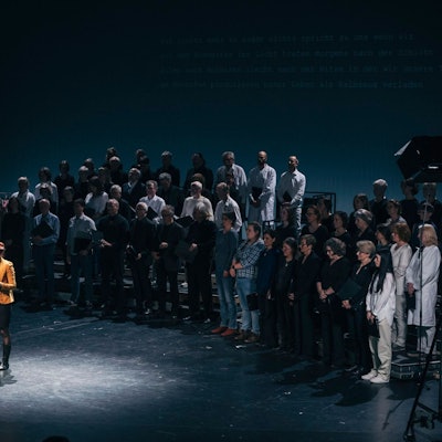 Daniela Danz bei der Poetica im Schauspiel Köln. Sie steht auf der Bühne, ein Lichtkegel hebt sie aus der Dunkelheit um sie herum heraus. Im Hintergrund ein Chor.