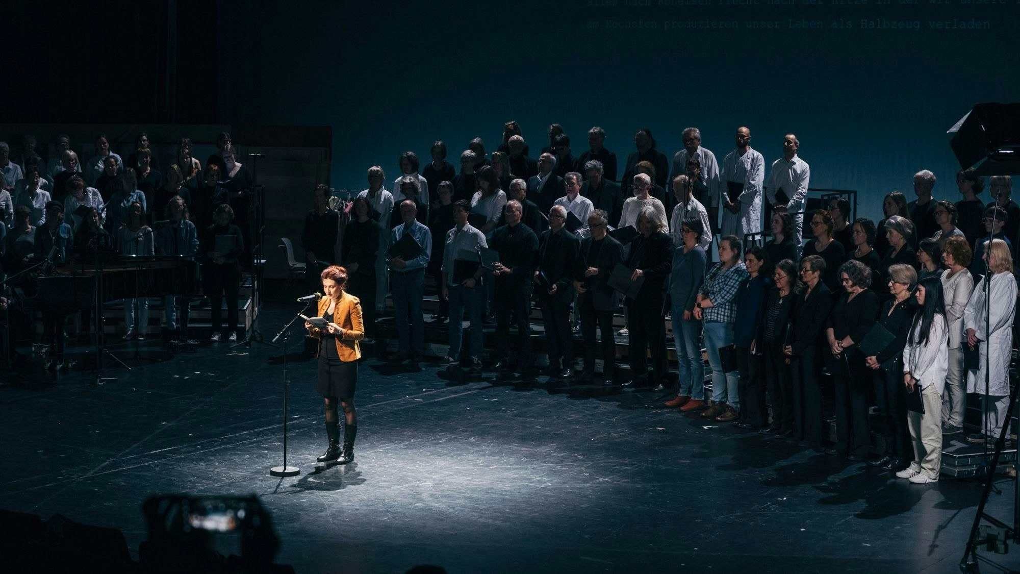 Daniela Danz bei der Poetica im Schauspiel Köln. Sie steht auf der Bühne, ein Lichtkegel hebt sie aus der Dunkelheit um sie herum heraus. Im Hintergrund ein Chor.