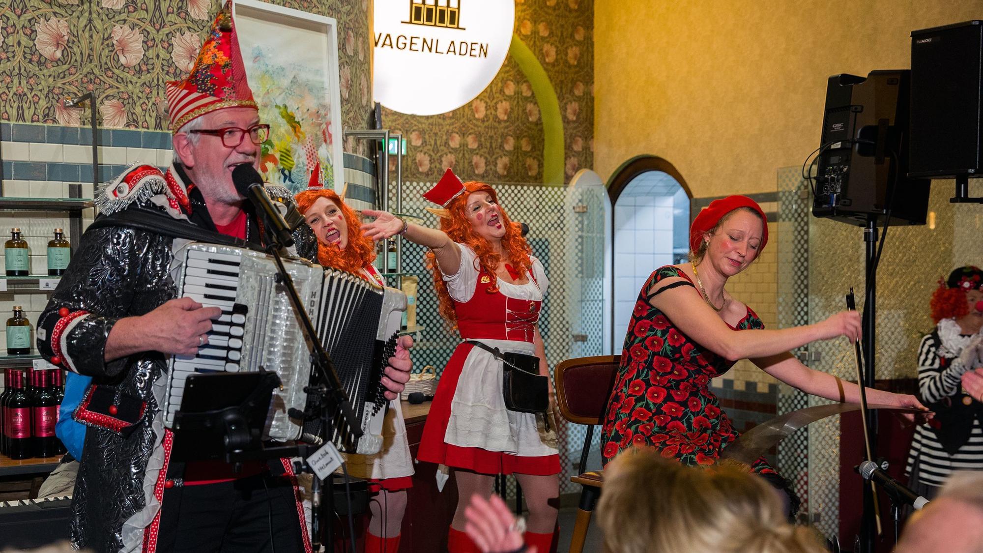 Ein verkleideter Mann, der Akkordeon spielt und in ein Mikrofon singt, zwei singende Frauen in einem Dirndl und eine Frau in einem Blumenkleid und Baskenmütze, die ein Instrument spielt
