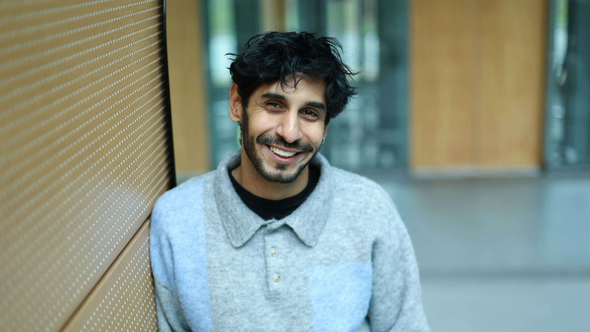 Juri Rother lehnt an einer Wand im Neven DuMont Haus. Er trägt einen grau-blauen Wollpullover und lacht in die Kamera.