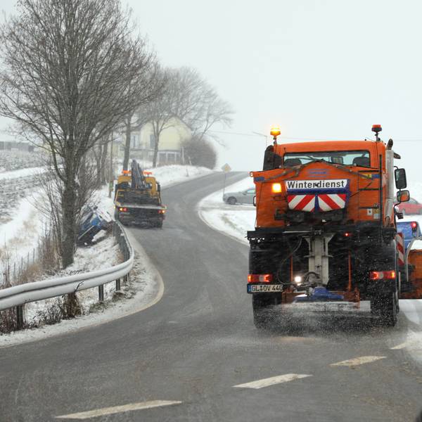 Ein Räumfahrzeug des Winterdienstes befreit eine Straße in der Eifel von Schneemassen. Weitere Schneeflocken fallen vom Himmel. (Symbolbild)