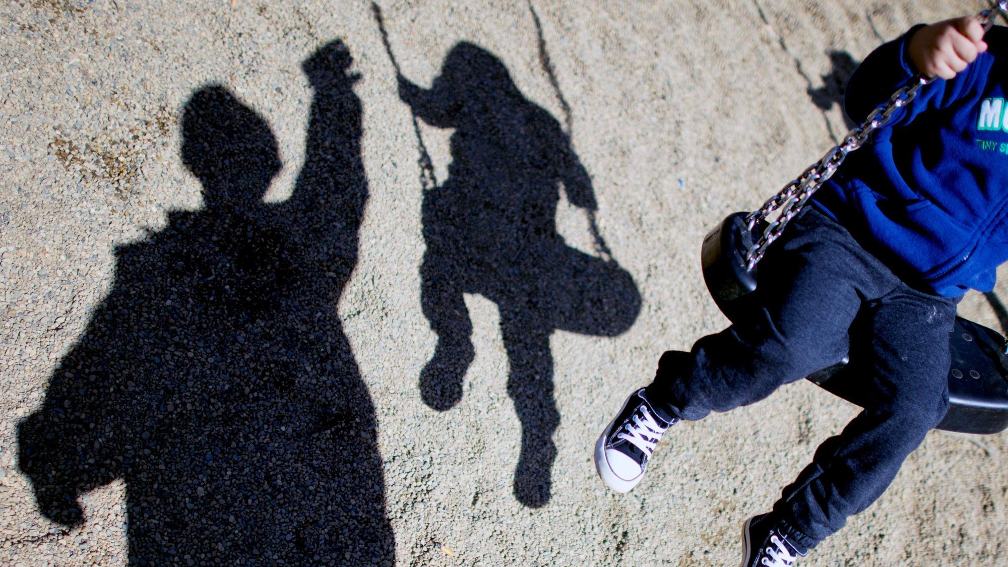 Zu sehen ist ein Kind auf einer Schaukel und sein Schatten, daneben ein weiterer Schatten eines Erwachsenen.