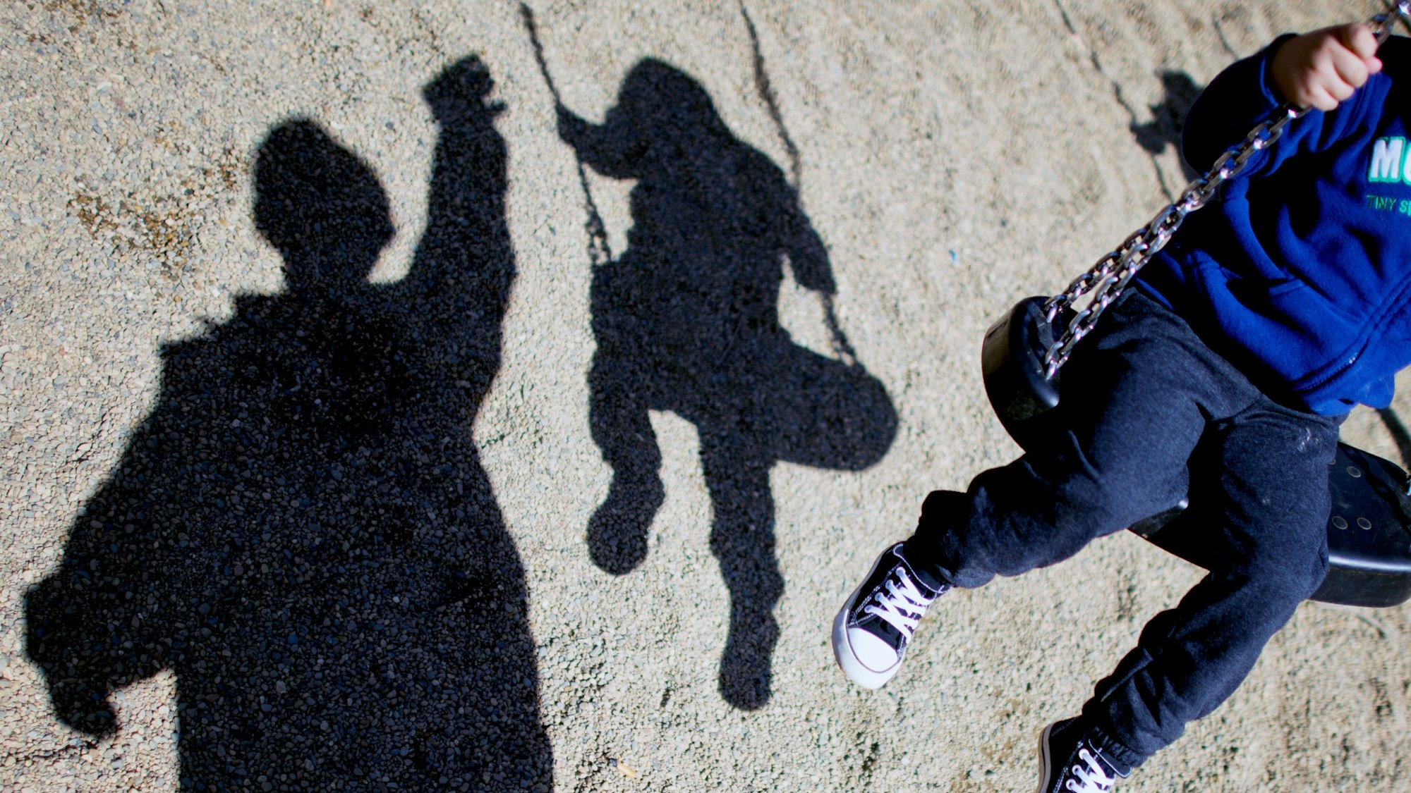 Zu sehen ist ein Kind auf einer Schaukel und sein Schatten, daneben ein weiterer Schatten eines Erwachsenen.