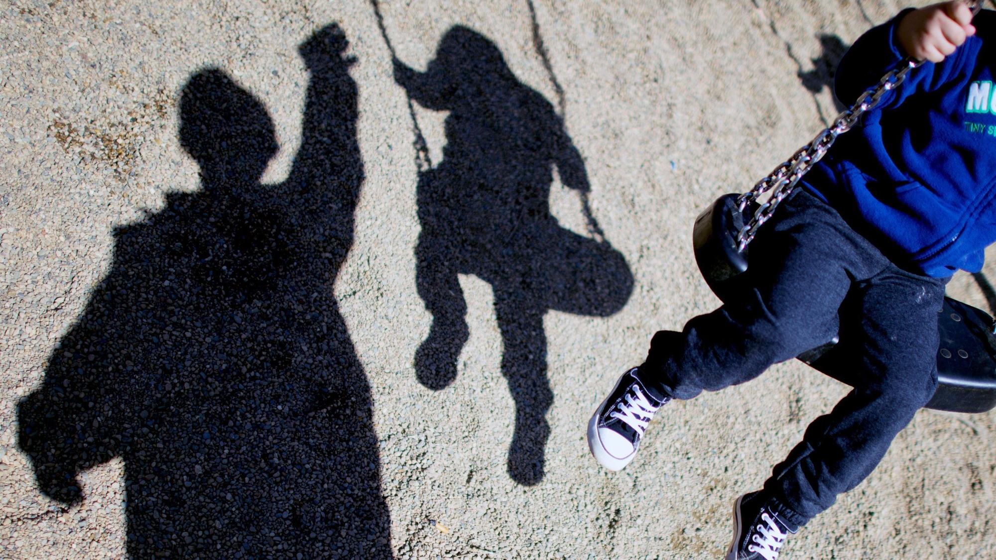Ein Kind sitzt auf einer Schaukel, sein Schatten ist auf dem Boden zu sehen, ebenso der Schatten einer weiteren Person.