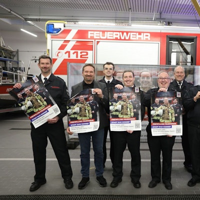 Eine Gruppe von acht Männern steht vor einem Feuerwehrfahrzeug. Sie halten Plakate in die Kamera.