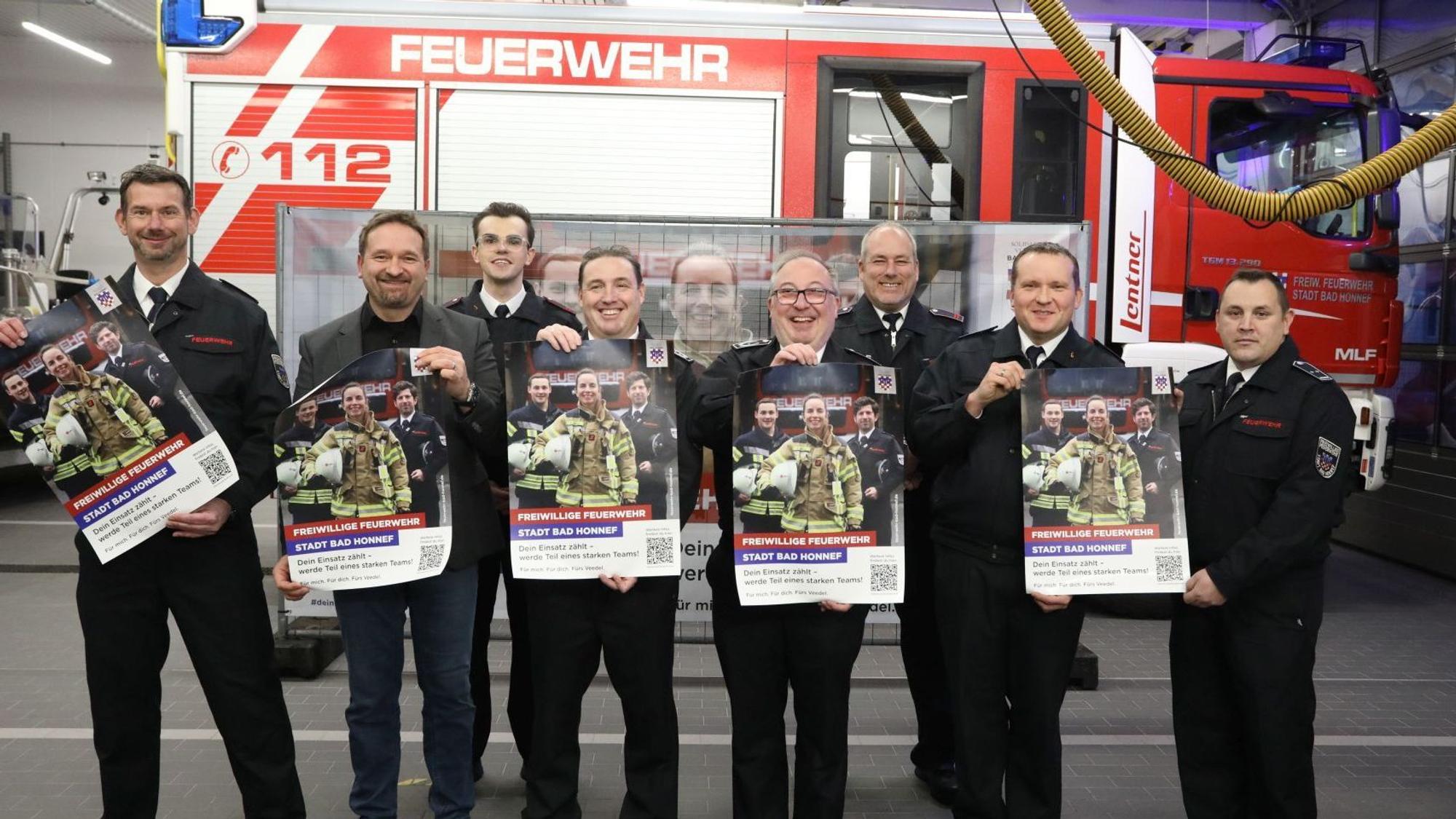 Eine Gruppe von acht Männern steht vor einem Feuerwehrfahrzeug. Sie halten Plakate in die Kamera.