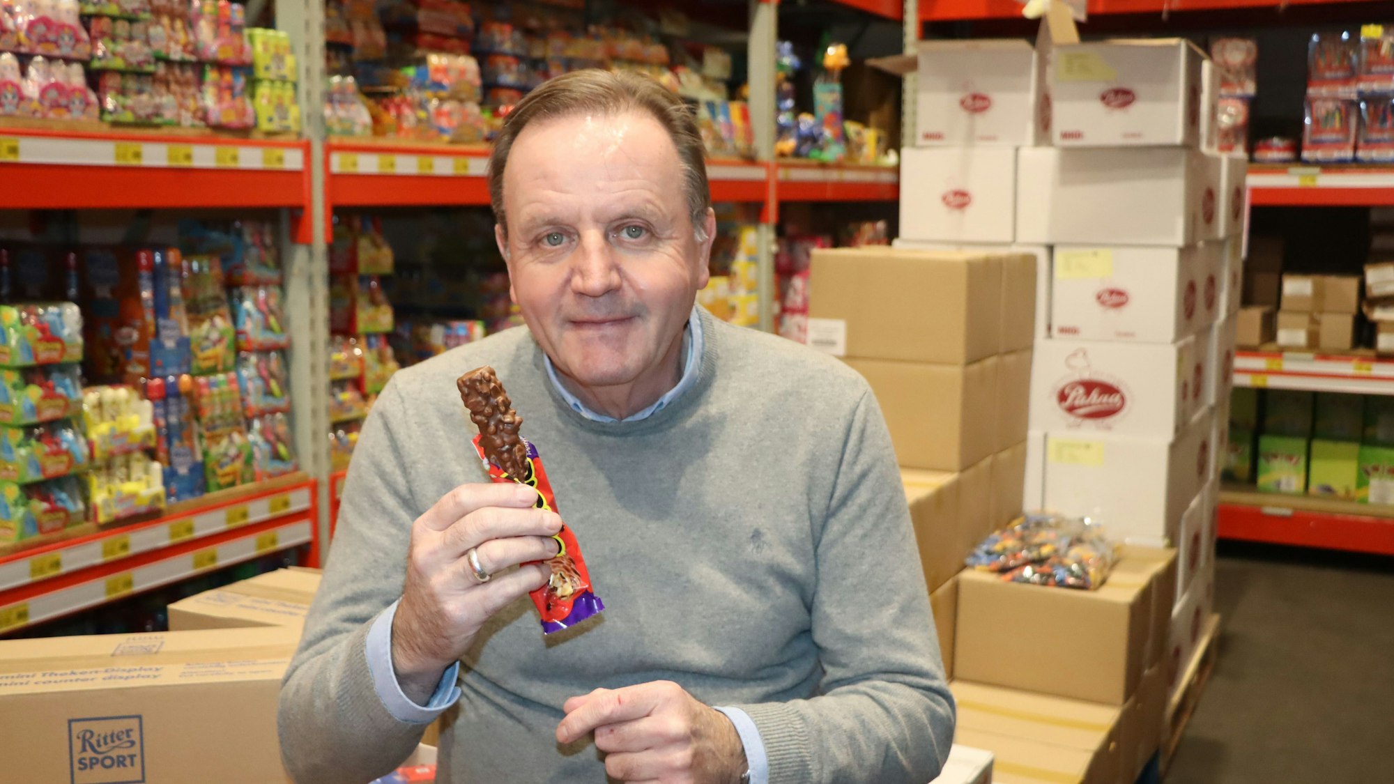 Ein Mann sitzt mit einem Schokoriegel vor Regalen voller Süßigkeiten.