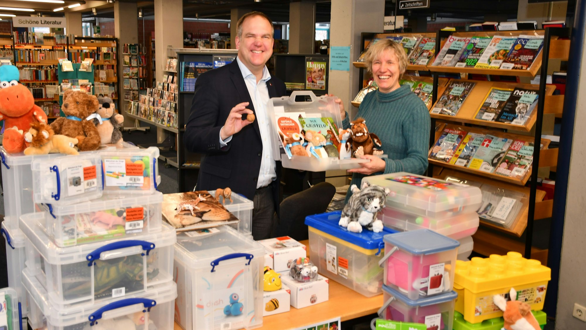 Der Bürgermeister und die Büchereileiterin zeigen einen Lesekoffer hinter einem Tisch mit Materialkisten in der Bücherei.