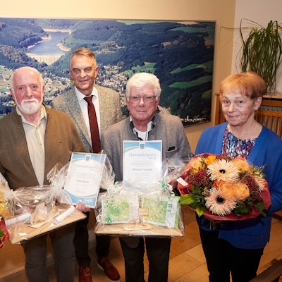 Drei Männer und zwei Frauen stehen vor einem Luftbild von Hellenthal. Die Frauen halten Blumen in den Händen, zwei der Männer Geschenke.