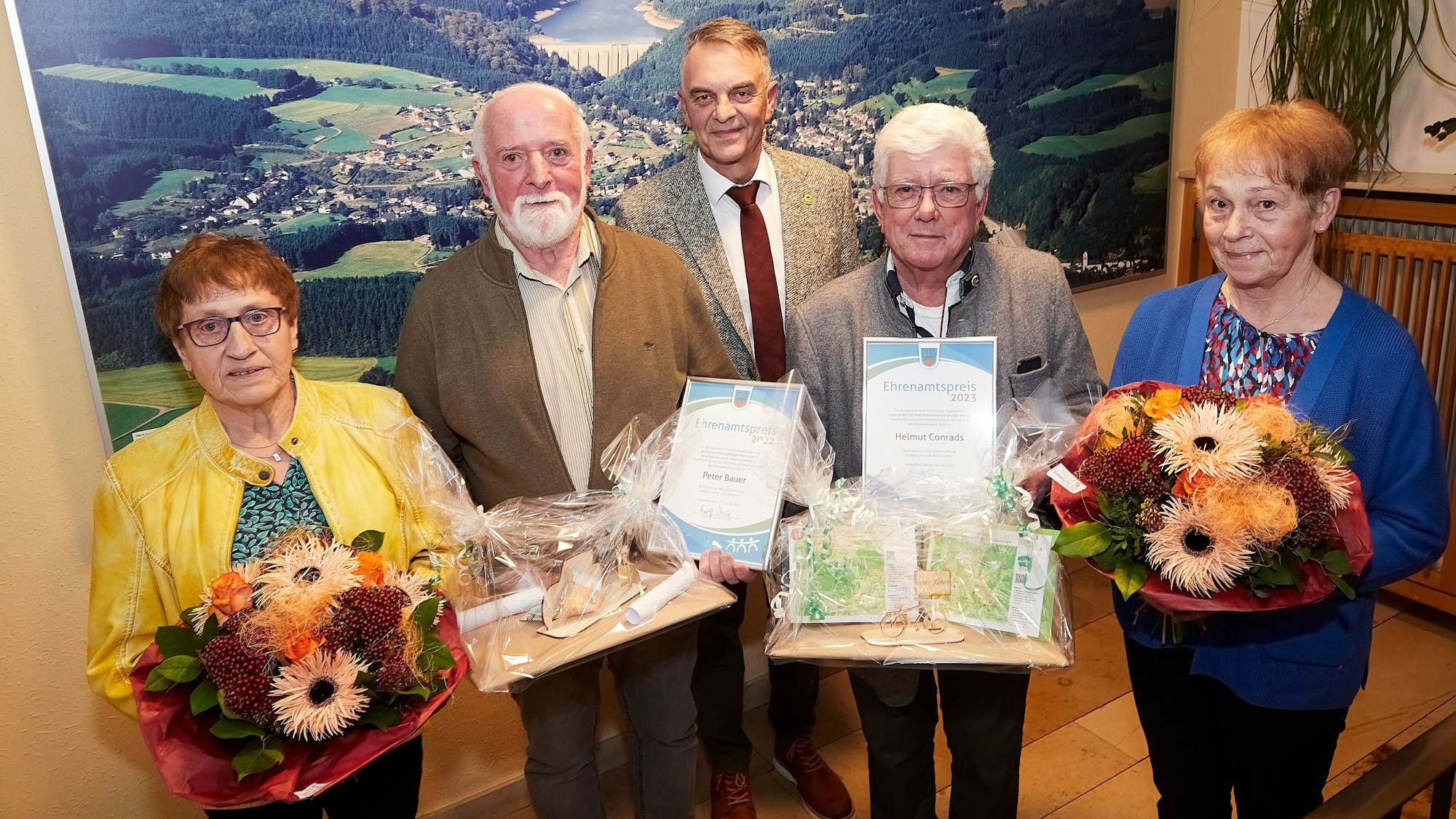Drei Männer und zwei Frauen stehen vor einem Luftbild von Hellenthal. Die Frauen halten Blumen in den Händen, zwei der Männer Geschenke.
