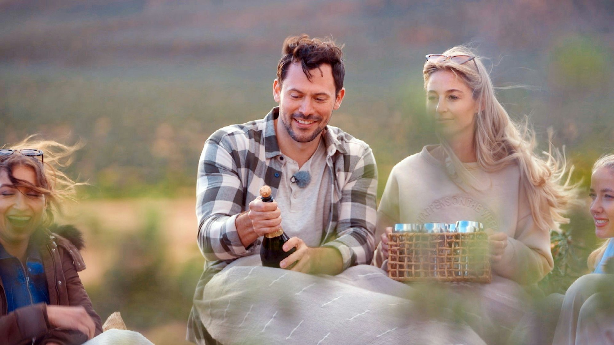 Ein Mann öffnet eine Sektflasche. Rechts und links neben ihm sitzen Frauen.