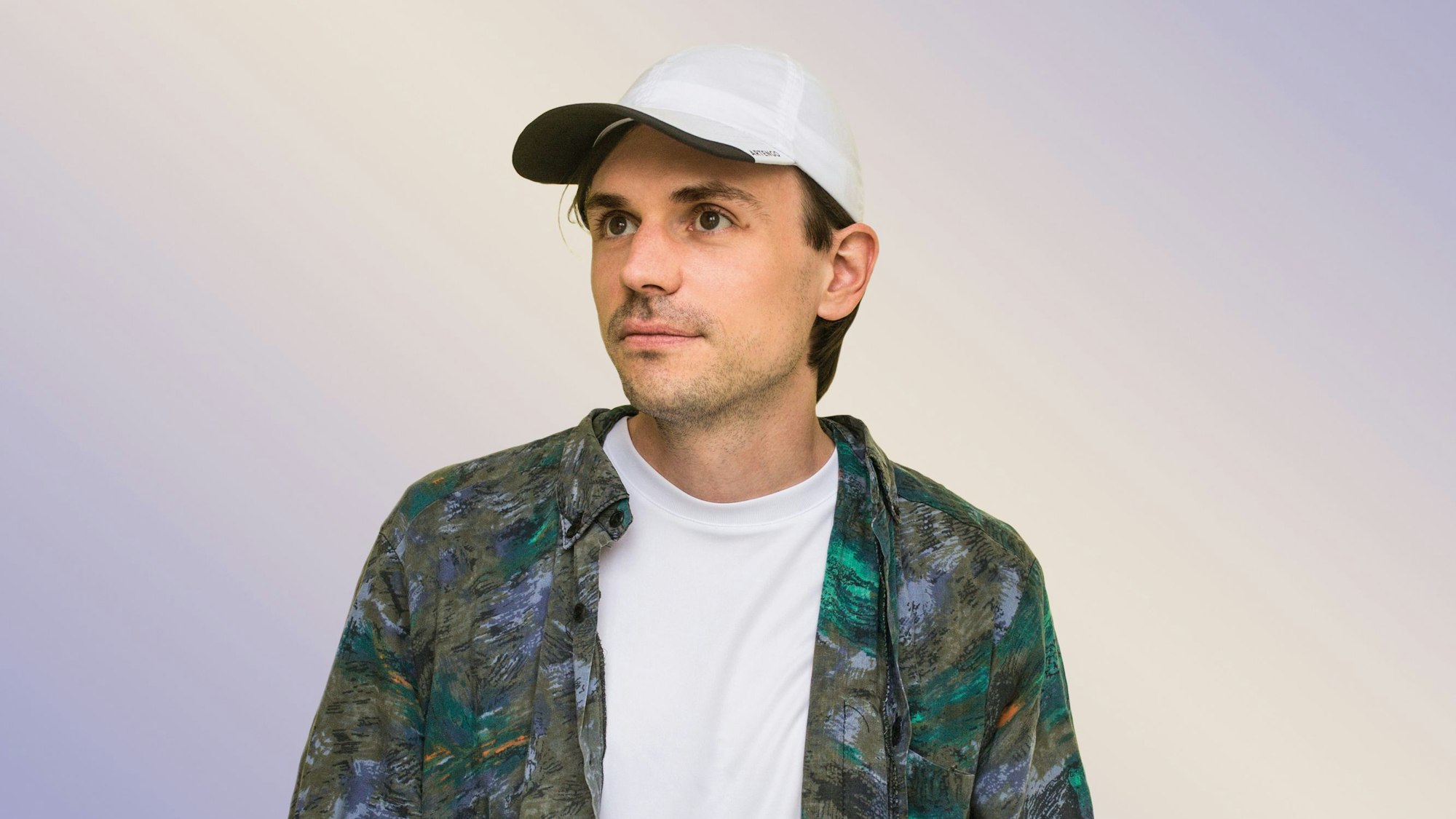 Leif Randt trägt ein buntes Hemd, ein weißes T-Shirt und eine weiße Mütze. Der Hintergrund ist pastellig-lila und beige.