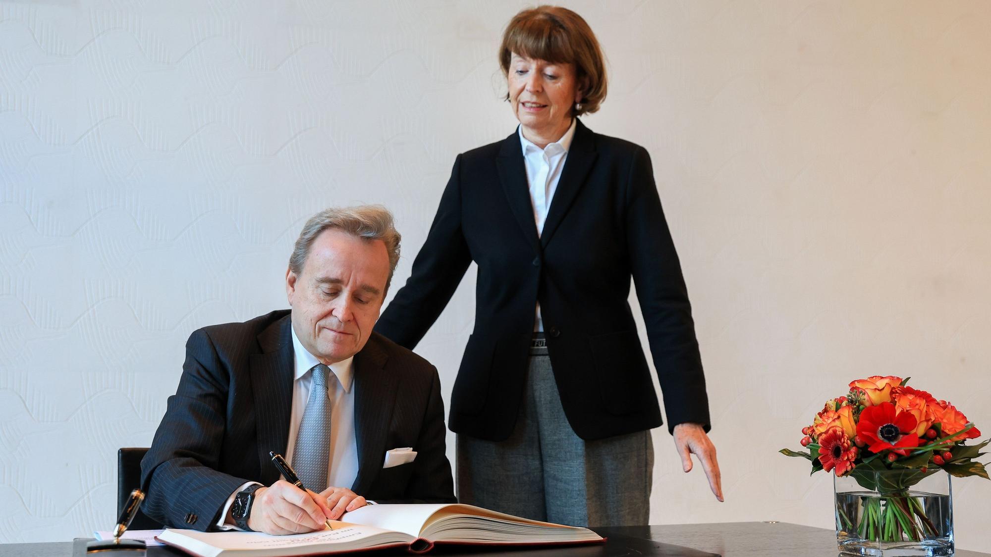 Bern Petelkau sitzt an einem Tisch und schreibt ins Gästebuch, Henriette Reker steht neben ihm.
