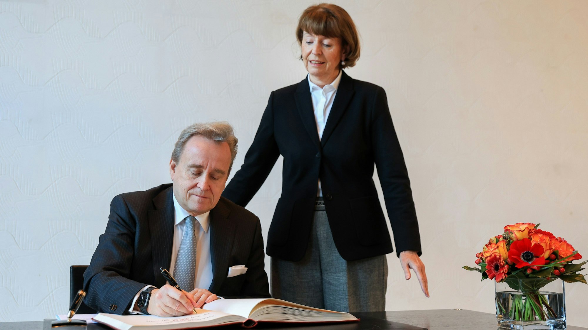 Bern Petelkau sitzt an einem Tisch und schreibt ins Gästebuch, Henriette Reker steht neben ihm.