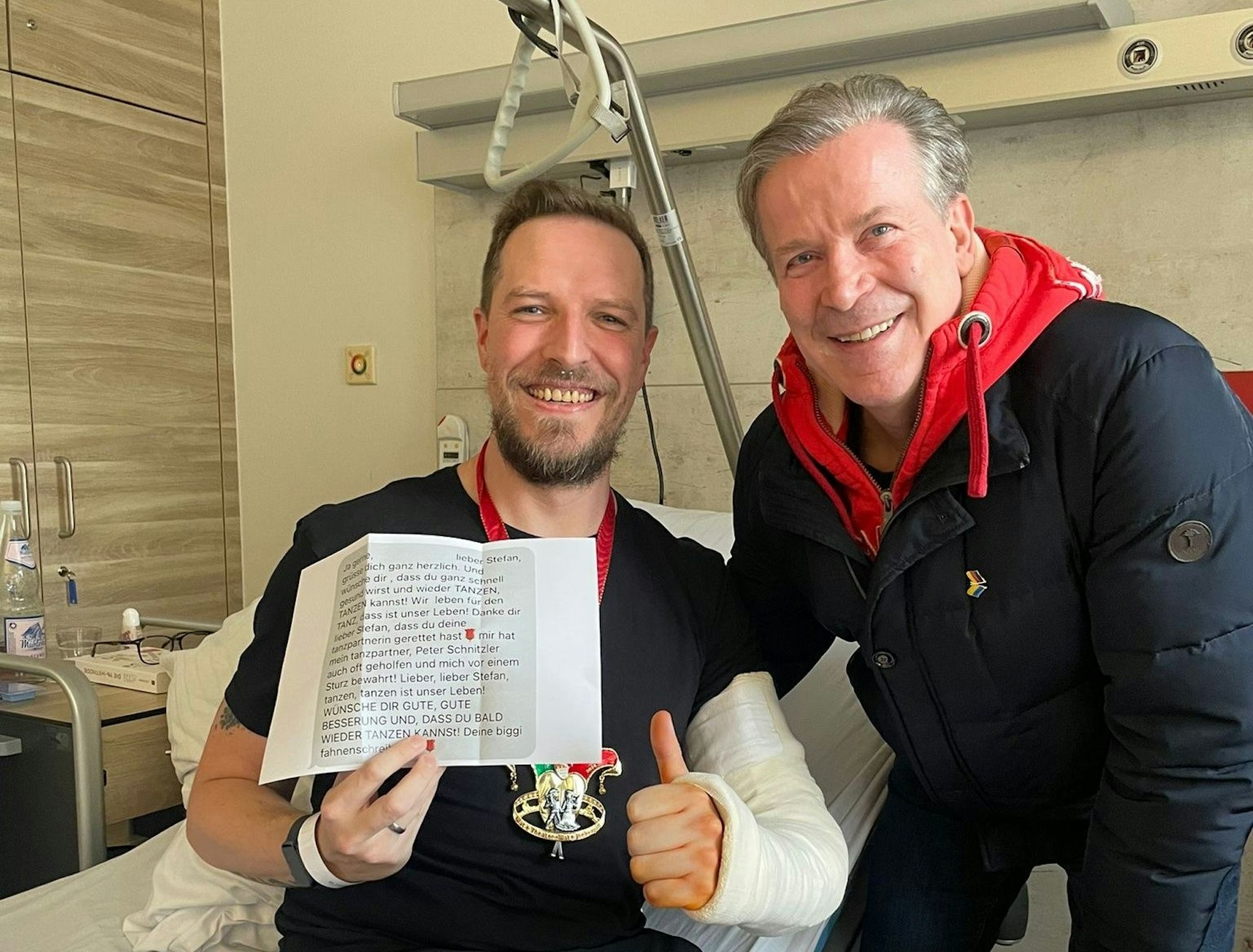 Zwei Männer lachen in die Kamera, einer trägt einen Gipsarm.
