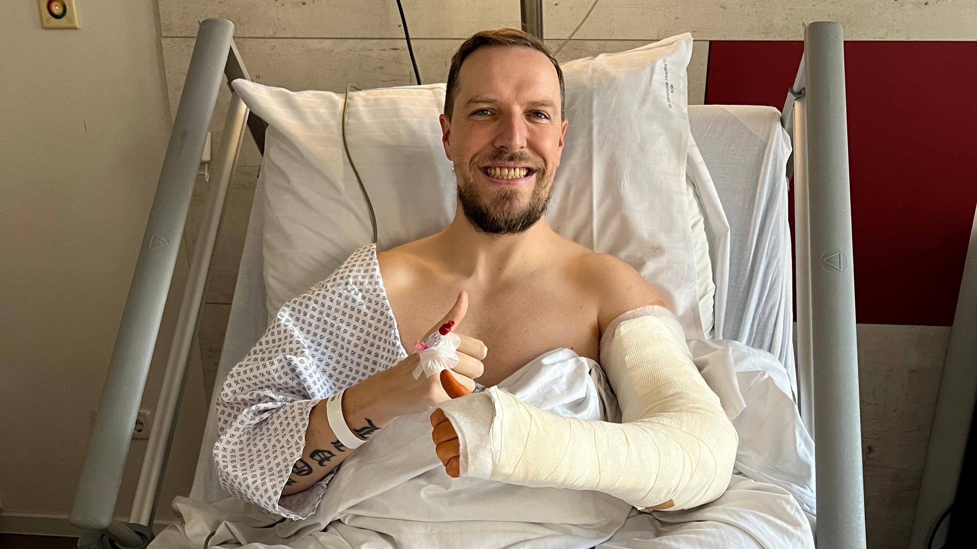 Ein Mann mit Gipsarm liegt auf einem Krankenhausbett und lacht in die Kamera-.