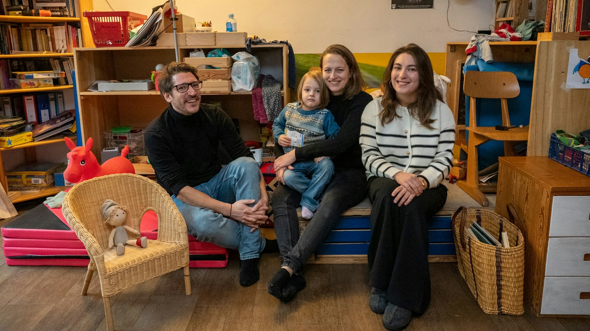 Zwei Frauen, ein Mann und ein kleines Kind sitzen in einem Wohnraum und schauen in die Kamera.