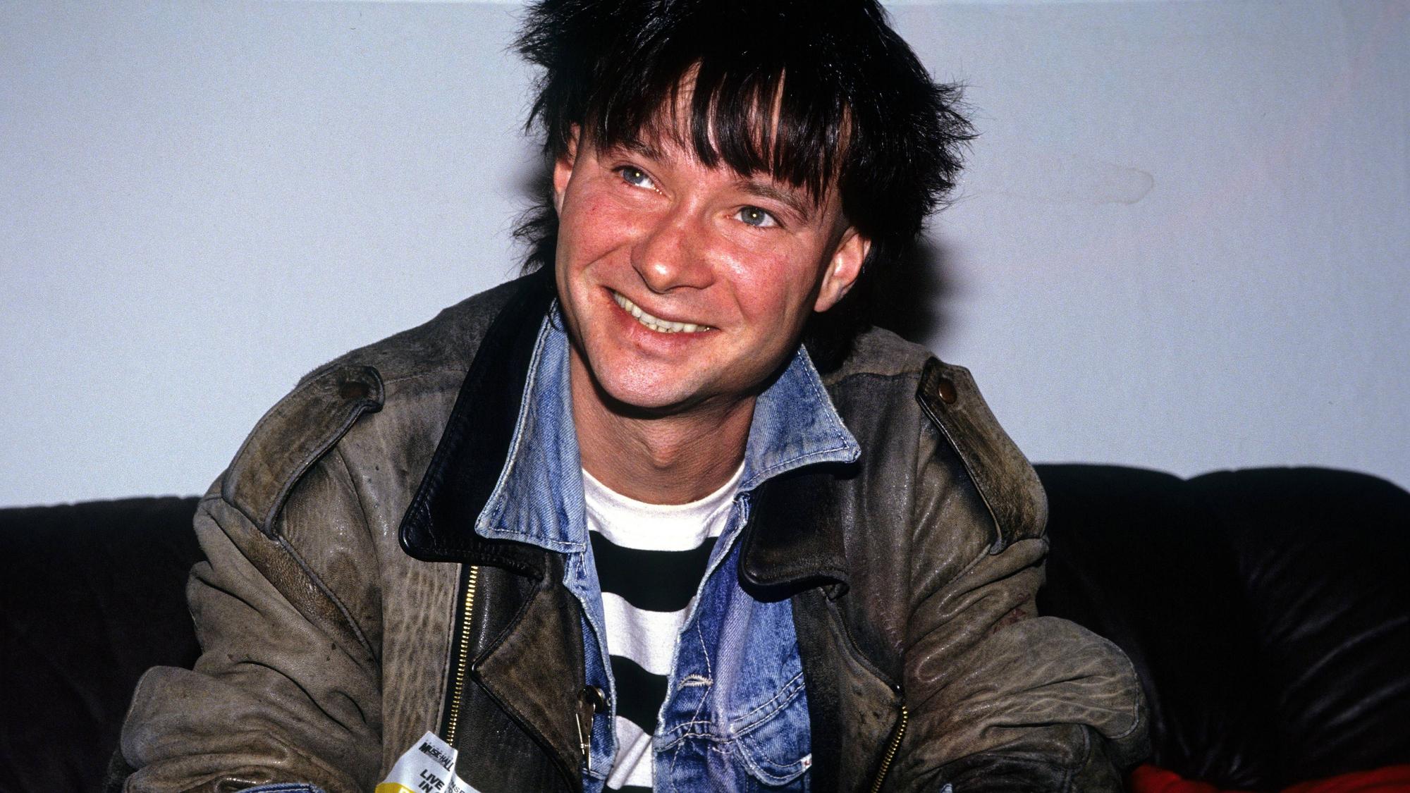 Frank Ziegert von der Hamburger Band Abwärts sitzt auf einem schwarzen Sofa. Er trägt eine alte Lederjacke über einer Jeansjacke, seine Haare sind schwarzgefärbt.