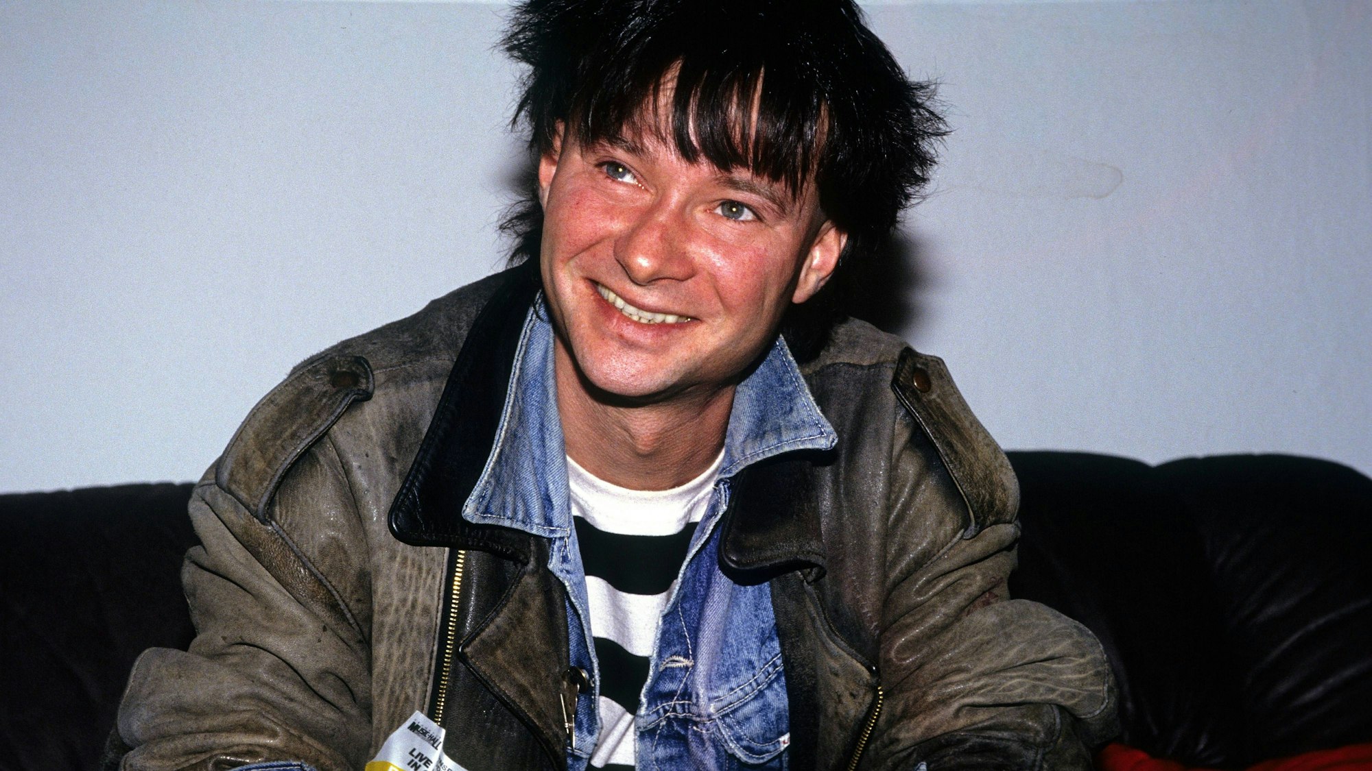 Frank Ziegert von der Hamburger Band Abwärts sitzt auf einem schwarzen Sofa. Er trägt eine alte Lederjacke über einer Jeansjacke, seine Haare sind schwarzgefärbt.