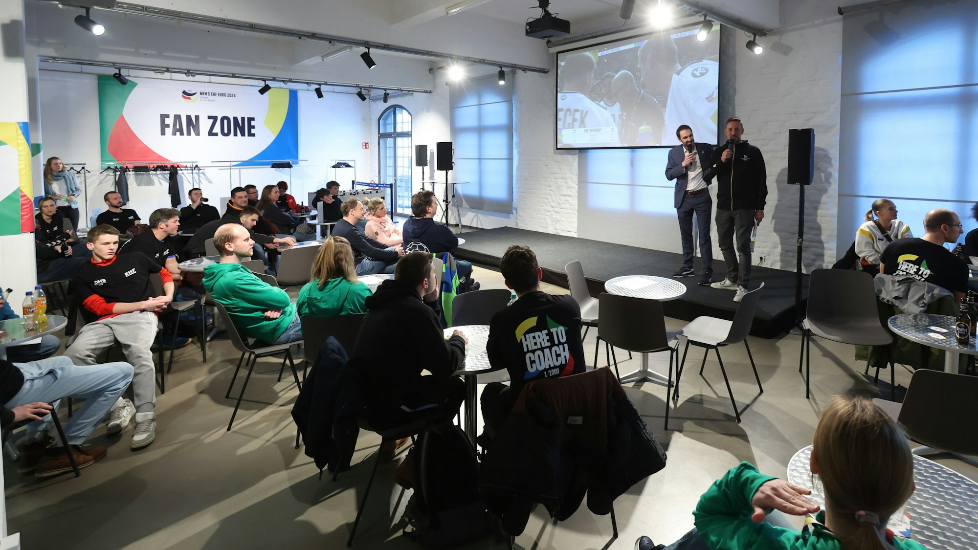 Auf dem Bild sieht man die Fan Zone im Deutschen Sport & Olympia Museum. Man sieht Personen, die das Spiel verfolgen.