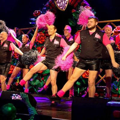 Männer in Cheerleader-Uniformen tanzen mit Pom-Poms.