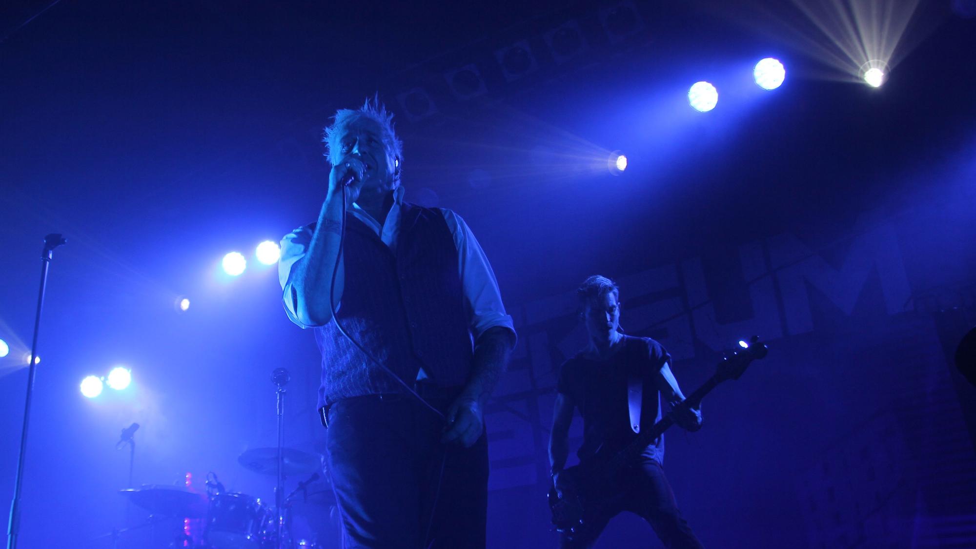 Auf einer in blaues Licht getauchten Bühne steht Sänger Michael Robert Rhein. Im Hintergrund sieht man Bassist Rupert Keplinger von der Band Eisbrecher.