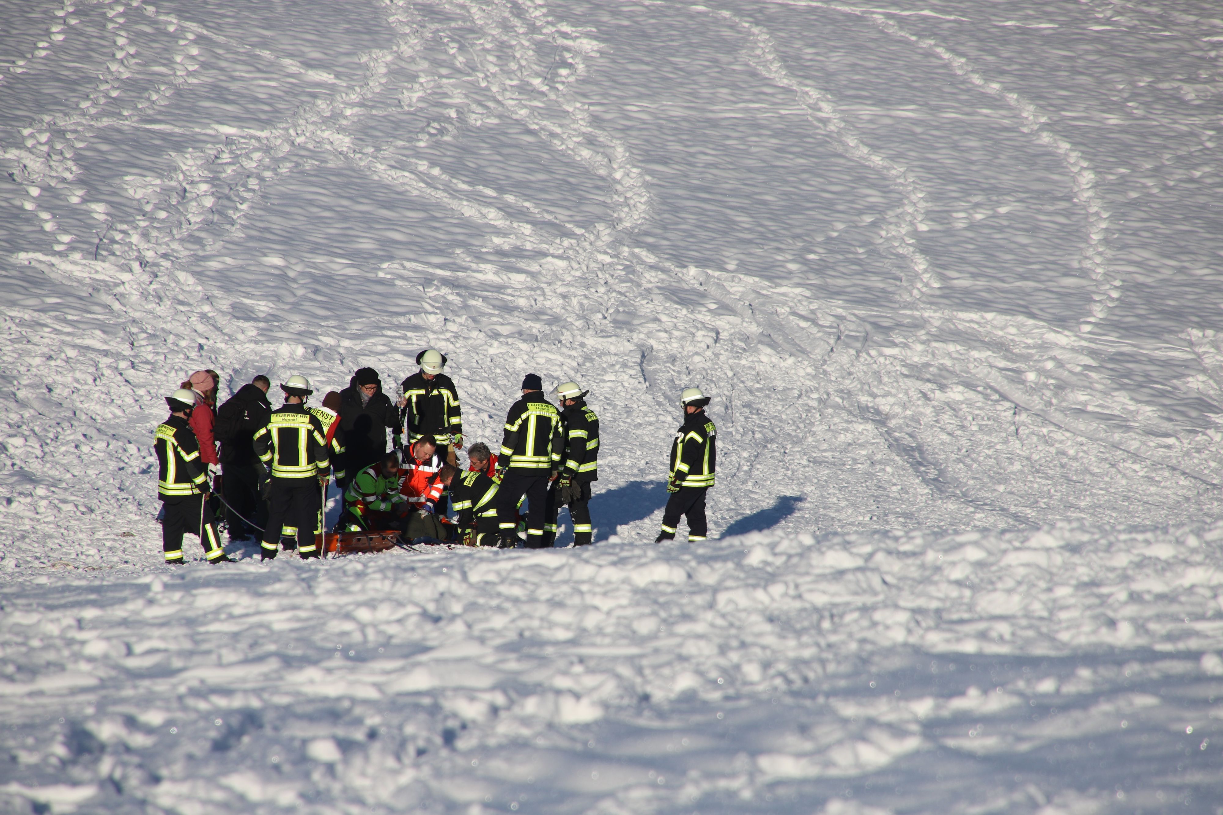 Rettungskräfte stehen auf einem tief verschneiten Hang. In ihrer Mitte knien Helfer und versorgen eine Patientin.