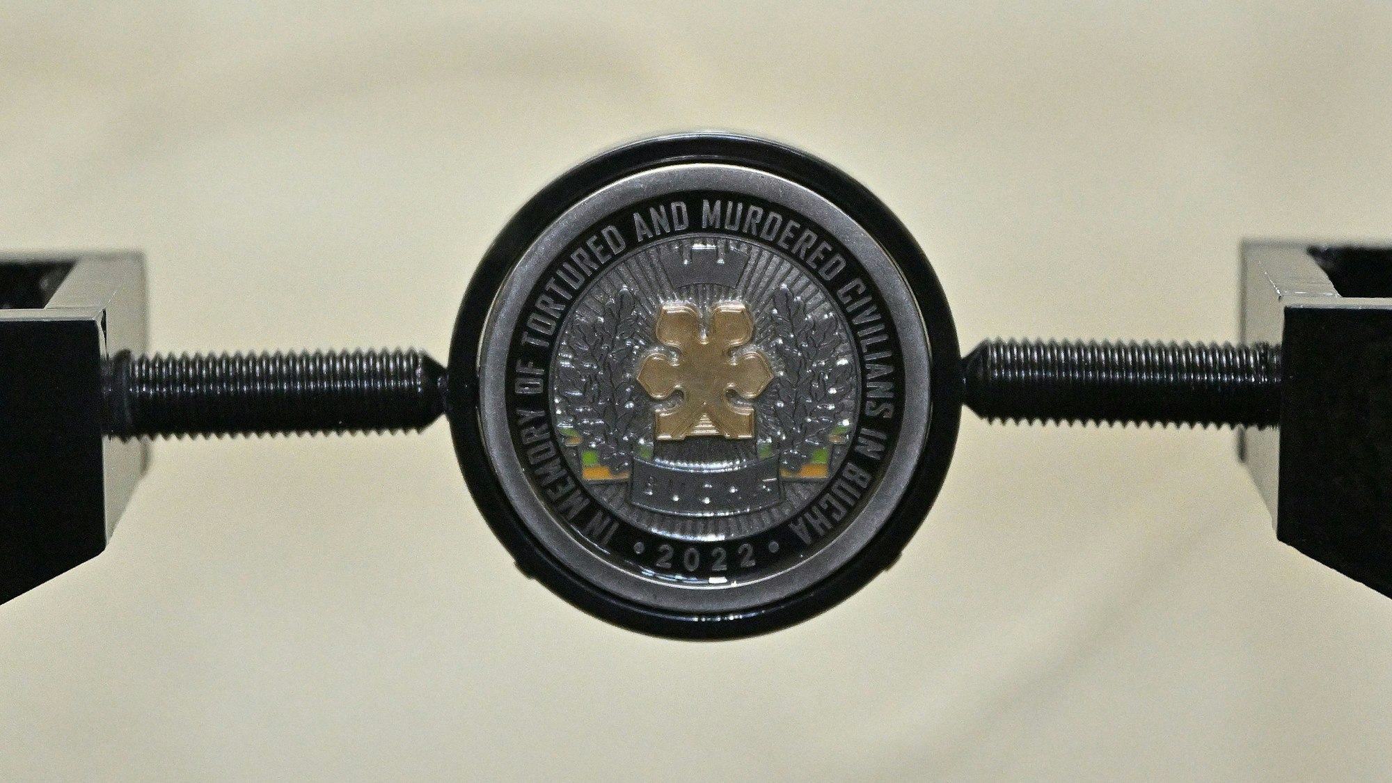 Eine Gedenkmünze an die Ermordeten in der ukrainischen Stadt Butscha ist in einer Halterung einer Stele befestigt.