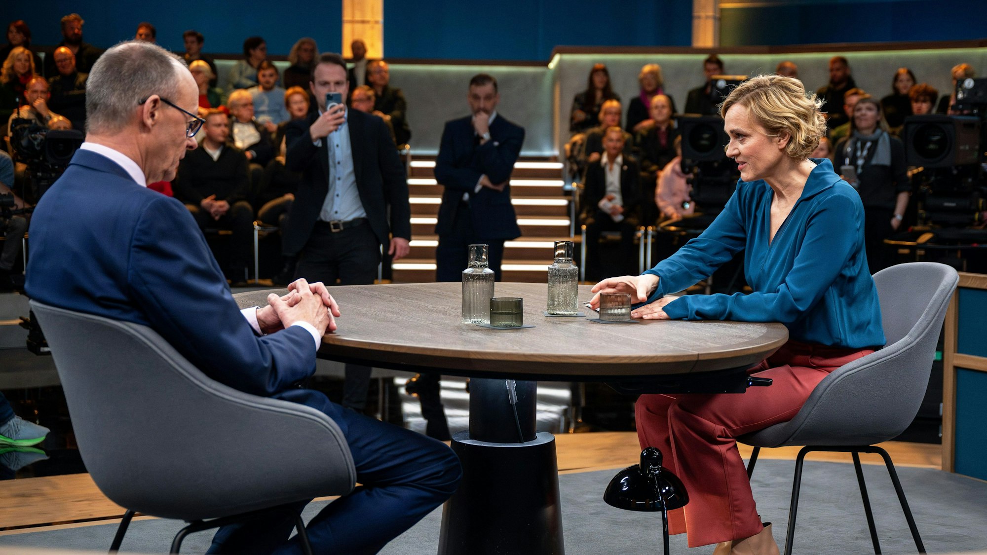 Caren Miosga sitzt mit Friedrich Merz im Studio ihres neuen Talks. Sie trägt eine blaue Bluse und eine rote Hose. Friedrich Merz trägt einen blauen Anzug