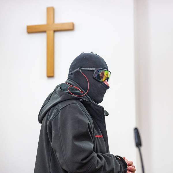 Ein Man in schwarzem Anorak, mit Sturmhaube und verspiegelter Sonnenbrille steht in einem Raum, an der Wand hängt ein Holzkreuz.