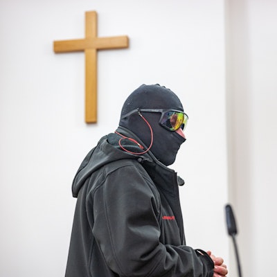 Ein Man in schwarzem Anorak, mit Sturmhaube und verspiegelter Sonnenbrille steht in einem Raum, an der Wand hängt ein Holzkreuz.