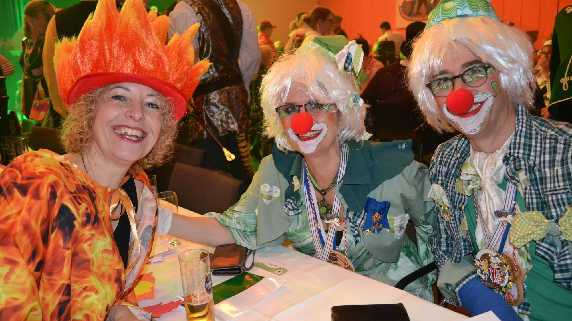 Drei lachende, bunt kostümierte Jecken sitzen an einem Tisch.