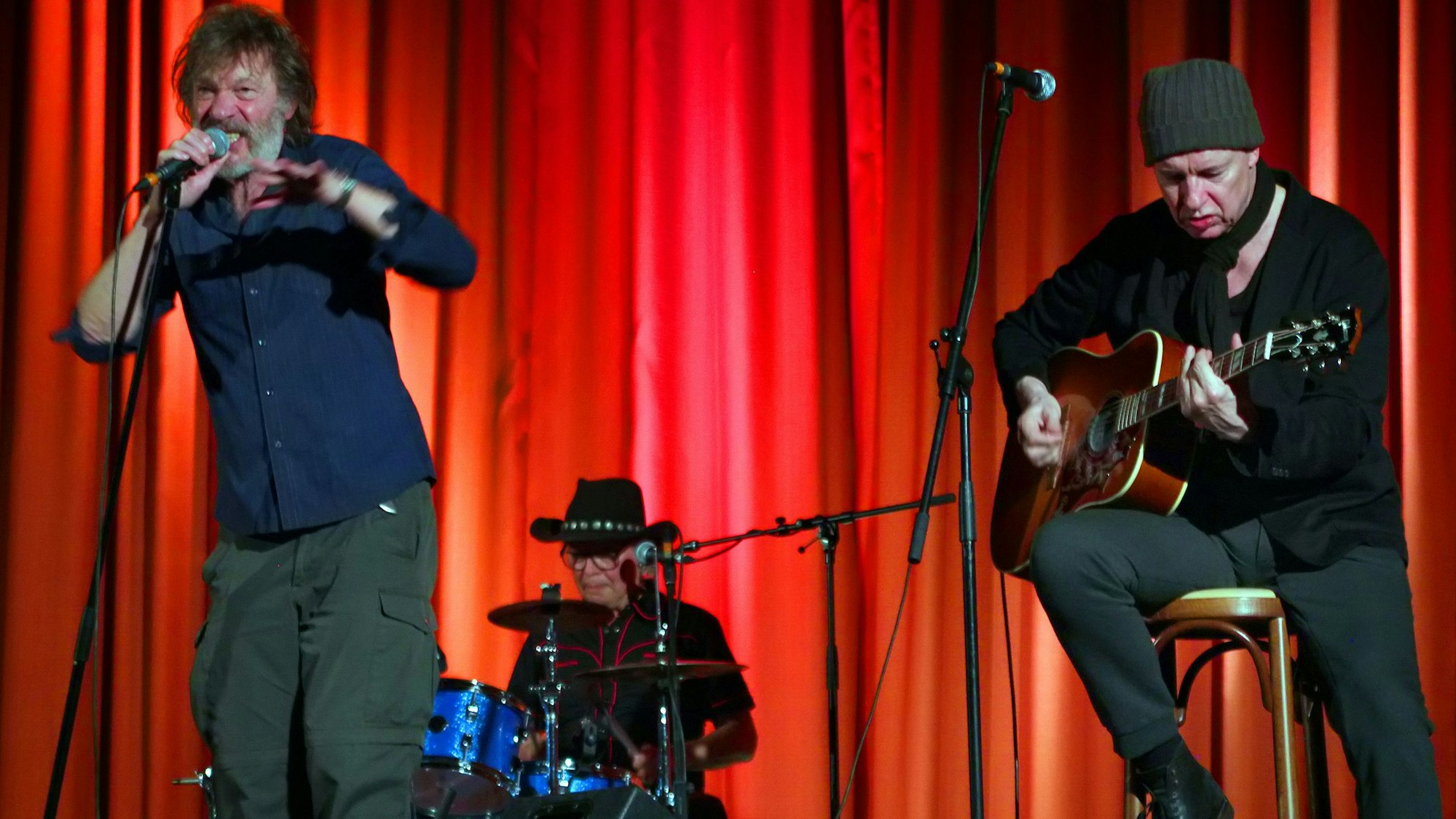 Ein Sänger, ein Schlagzeuger und ein Gitarrist auf einer Bühne vor einem roten Vorhang.