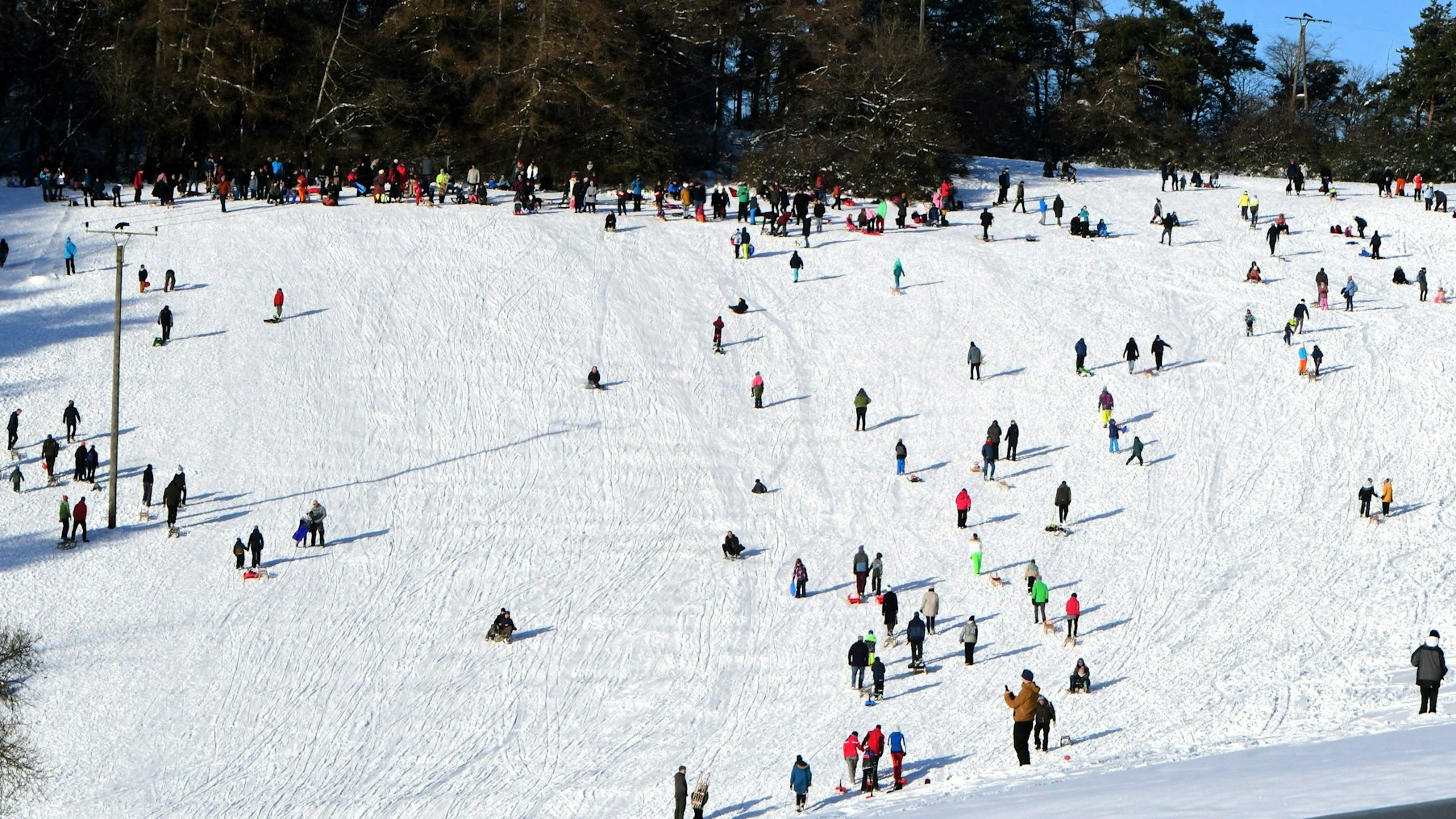 Zahlreiche Schlittenfahrer und Spaziergänger sind auf dem verschneiten Hang unterwegs.