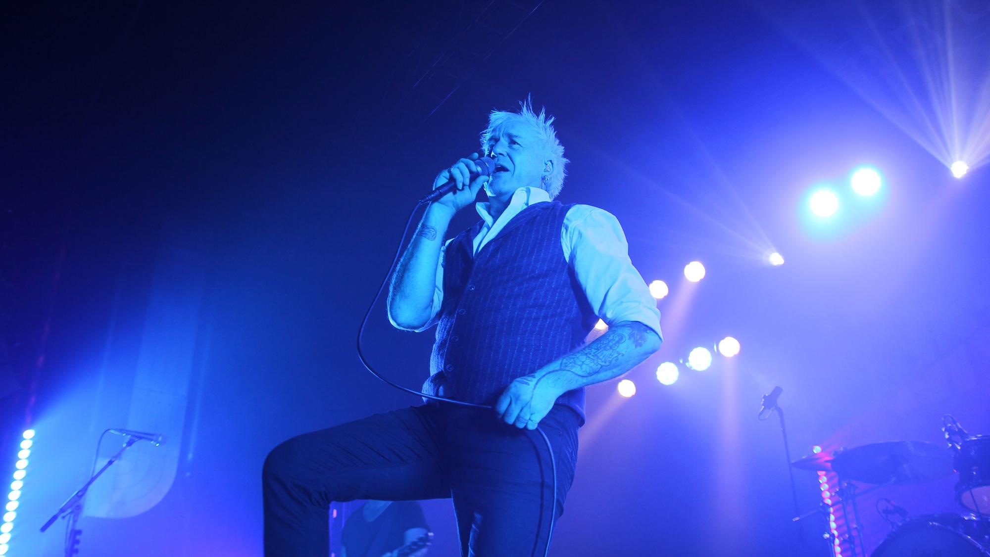 Sänger Michael Robert Rhein steht mit einem Mikrofon vor dem Mund auf der Bühne, die in blaues Licht getaucht ist.