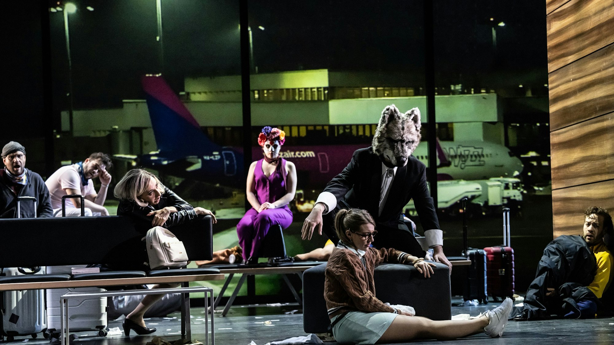 Auf der Bühne ist eine Szene in einem Flughafenterminal aufgebaut. Die Schauspieler sitzen wartend teils auch auf dem Boden. Einer trägt eine Wolfsmaske.