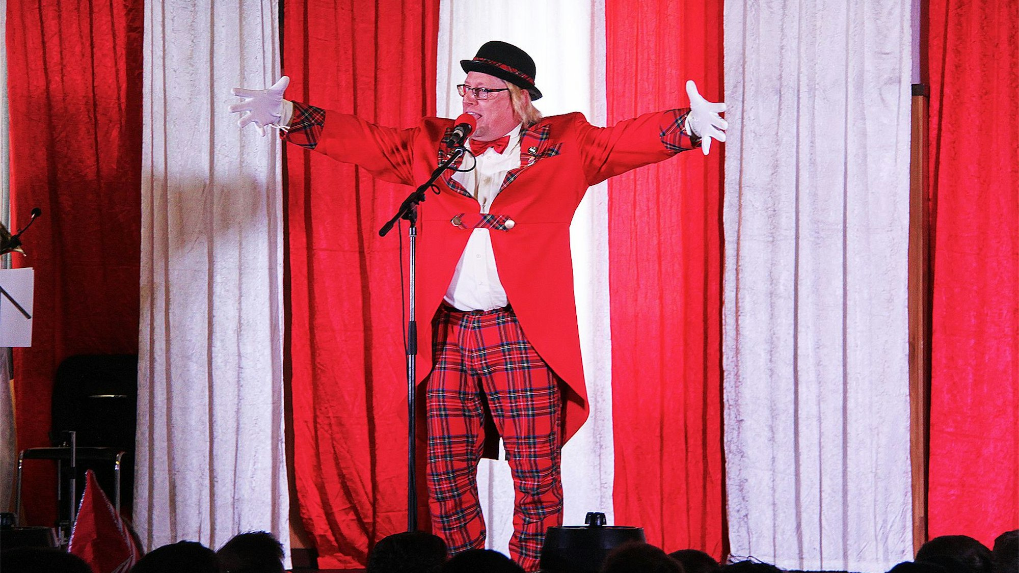 Der Redner Stefan van der Eerdwich steht gestikulierend auf der rot-weiß dekorierten Bühne. Er trägt einen schwarzen Hut, einen roten Frack und eine rot-schwarz karierte Hose.