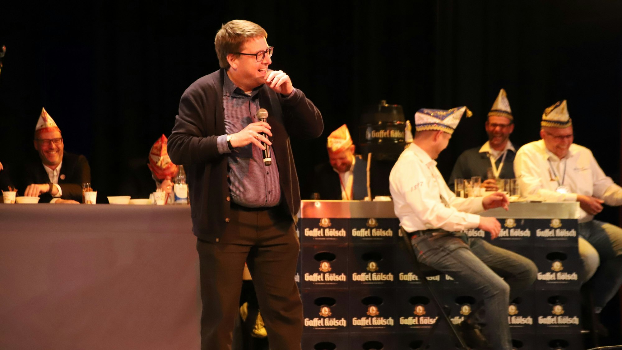 Ein Mann mit Mikrofon auf einer Bühne. Im Hintergrund sitzen Männer mit weißen Hemden und Karnevalskappen an einer Bar aus Bierkästen.