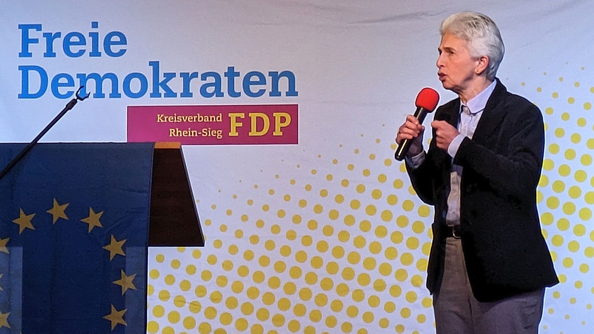 Eine Frau mit kurzen grauen Haaren, schwarzem Sakko und grauer Hose spricht in ein Mikrofon. Auf der Wand steht Freie Demokraten.