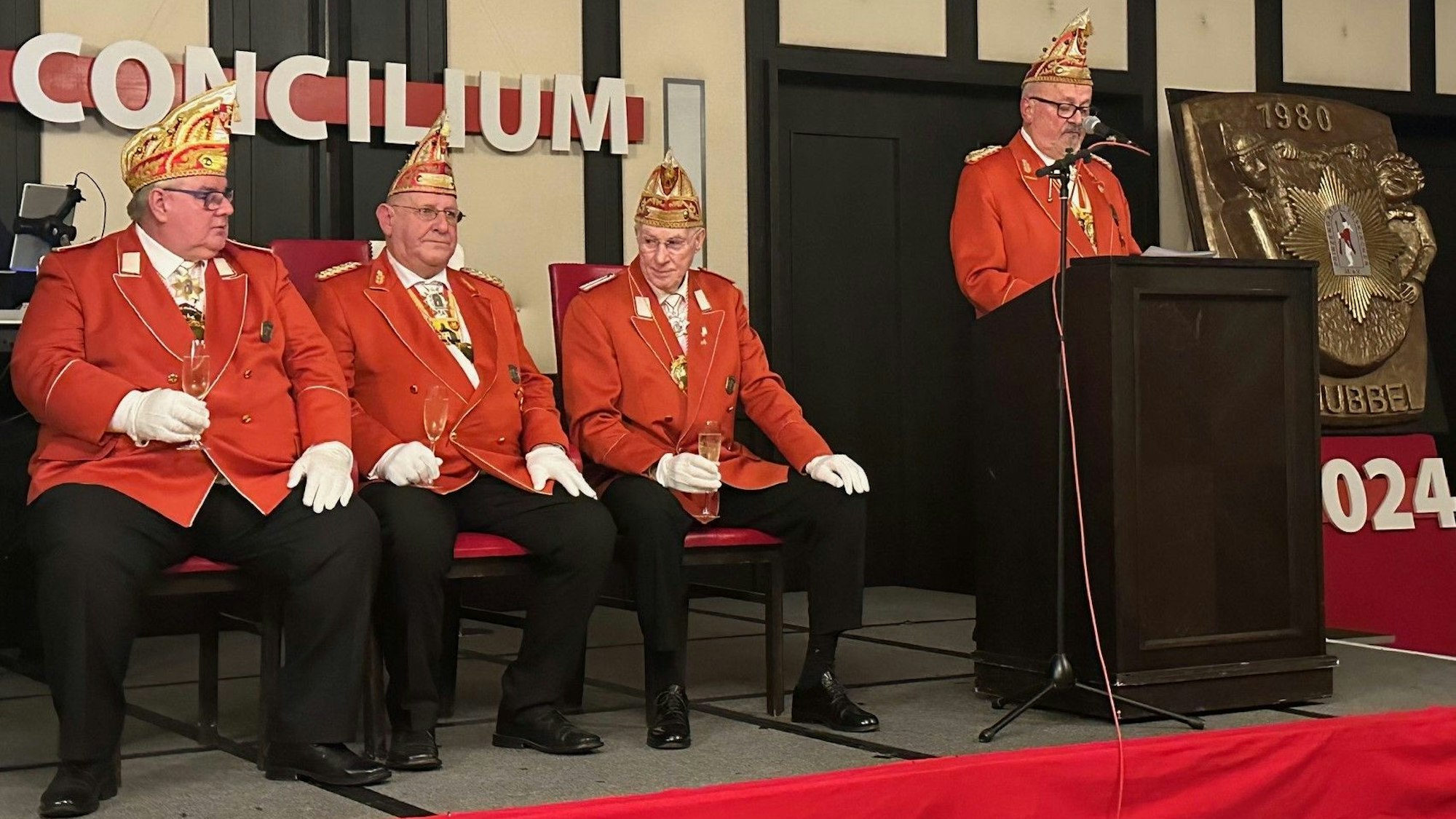 Flankiert von weiteren Karnevalisten, hört Klaus Peter Neuburg sitzend die Laudatio, die Ferdy Pfahl am Rednerpult hält.